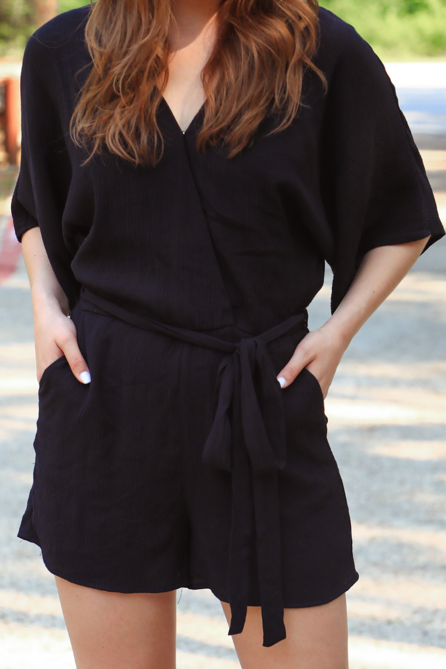 a black lined romper made of soft lightweight fabric with a surplice V-neckline, snap closure, relaxed flowy sleeves, waist-defining accent tie, side pockets, and a flattering easy-to-wear silhouette