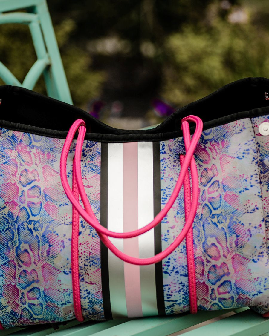 ultra-lightweight tote made of neoprene with a removable hard bottom liner, paracord shoulder straps, removable matching zippered pouch, magnetic side snap closures in silver hardware, black interior (tote and pouch), and racing stripes (black, gray, and pink) on a soft pastel snakeskin pattern