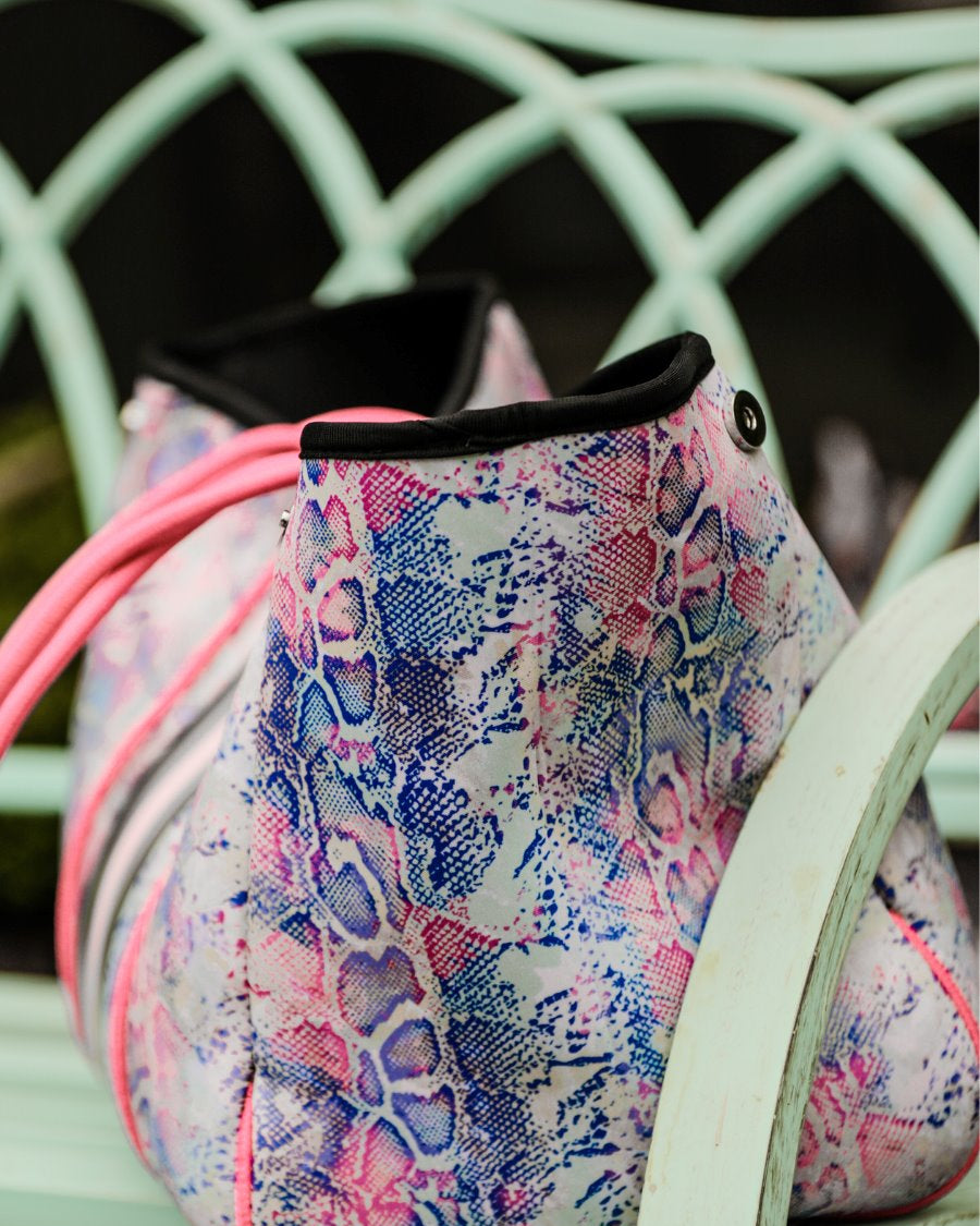 ultra-lightweight tote made of neoprene with a removable hard bottom liner, paracord shoulder straps, removable matching zippered pouch, magnetic side snap closures in silver hardware, black interior (tote and pouch), and racing stripes (black, gray, and pink) on a soft pastel snakeskin pattern