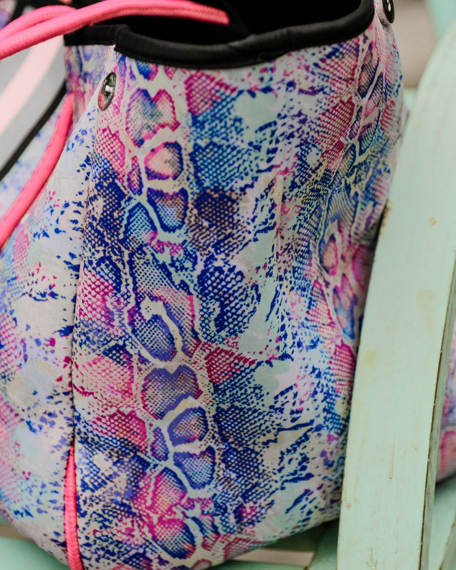 ultra-lightweight tote made of neoprene with a removable hard bottom liner, paracord shoulder straps, removable matching zippered pouch, magnetic side snap closures in silver hardware, black interior (tote and pouch), and racing stripes (black, gray, and pink) on a soft pastel snakeskin pattern