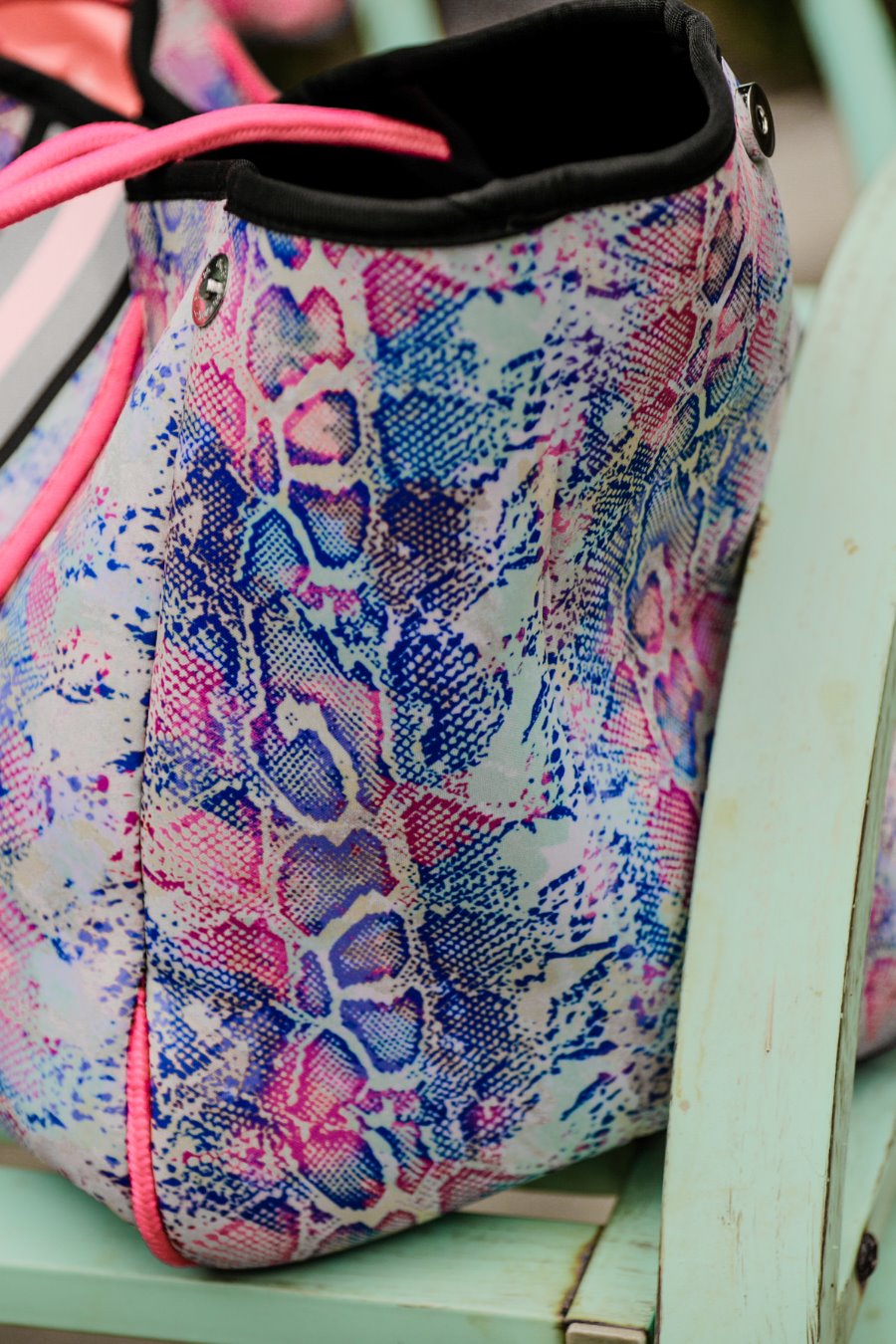 ultra-lightweight tote made of neoprene with a removable hard bottom liner, paracord shoulder straps, removable matching zippered pouch, magnetic side snap closures in silver hardware, black interior (tote and pouch), and racing stripes (black, gray, and pink) on a soft pastel snakeskin pattern