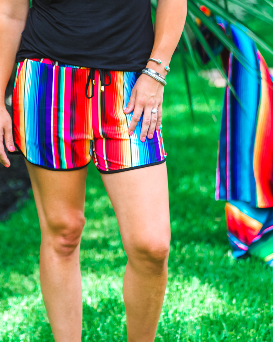 mid-rise relaxed fit activewear shorts with side pockets and a tie waist drawstring with gold bead accents, designed on a multicolor serape pattern print fabric and black trim