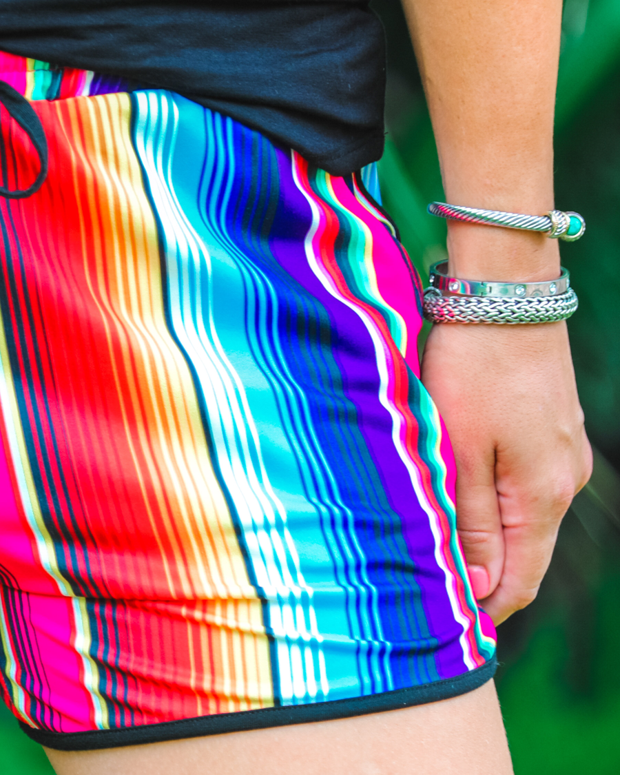 mid-rise relaxed fit activewear shorts with side pockets and a tie waist drawstring with gold bead accents, designed on a multicolor serape pattern print fabric and black trim