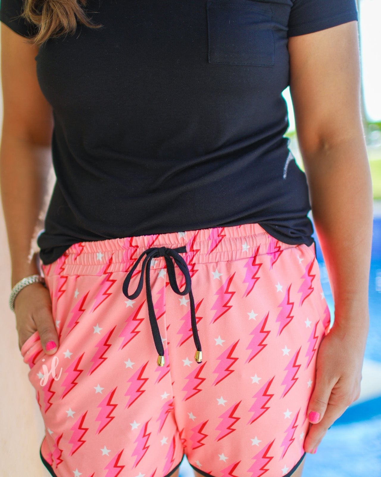 mid-rise relaxed fit activewear shorts with side pockets and a tie waist drawstring with gold bead accents, designed on a two-tone pink and red lightning bolts and white stars pattern print fabric, coral base, and black trim
