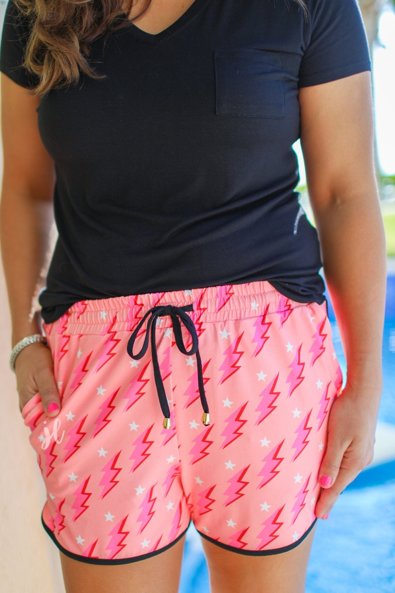 mid-rise relaxed fit activewear shorts with side pockets and a tie waist drawstring with gold bead accents, designed on a two-tone pink and red lightning bolts and white stars pattern print fabric, coral base, and black trim