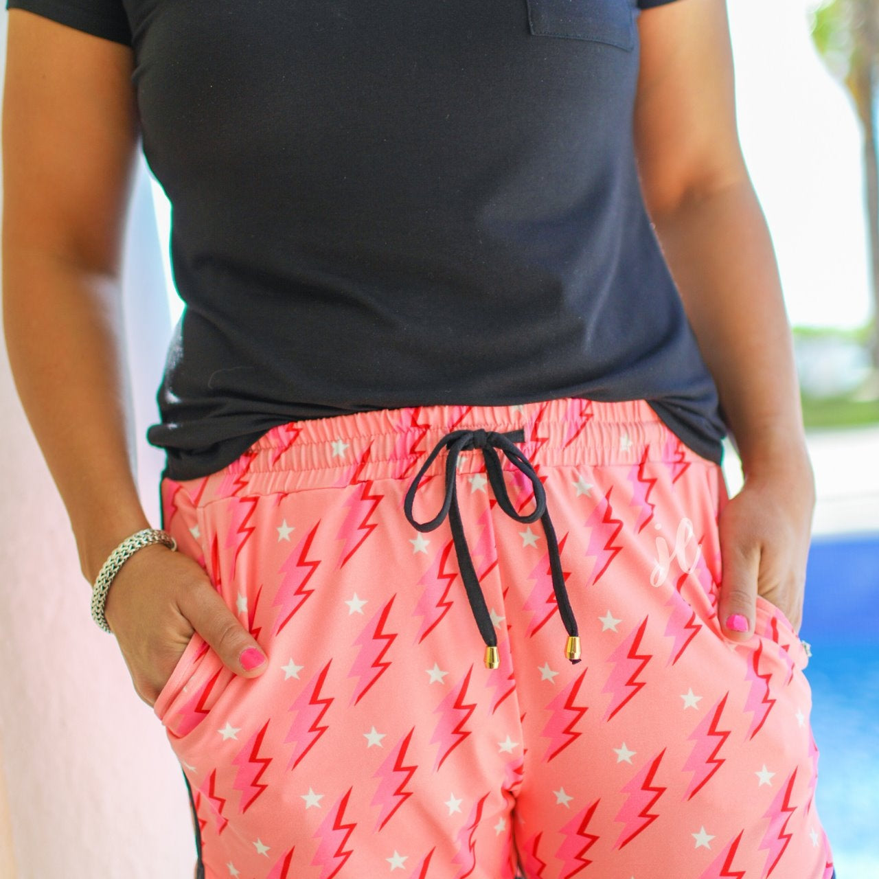 mid-rise relaxed fit activewear shorts with side pockets and a tie waist drawstring with gold bead accents, designed on a two-tone pink and red lightning bolts and white stars pattern print fabric, coral base, and black trim