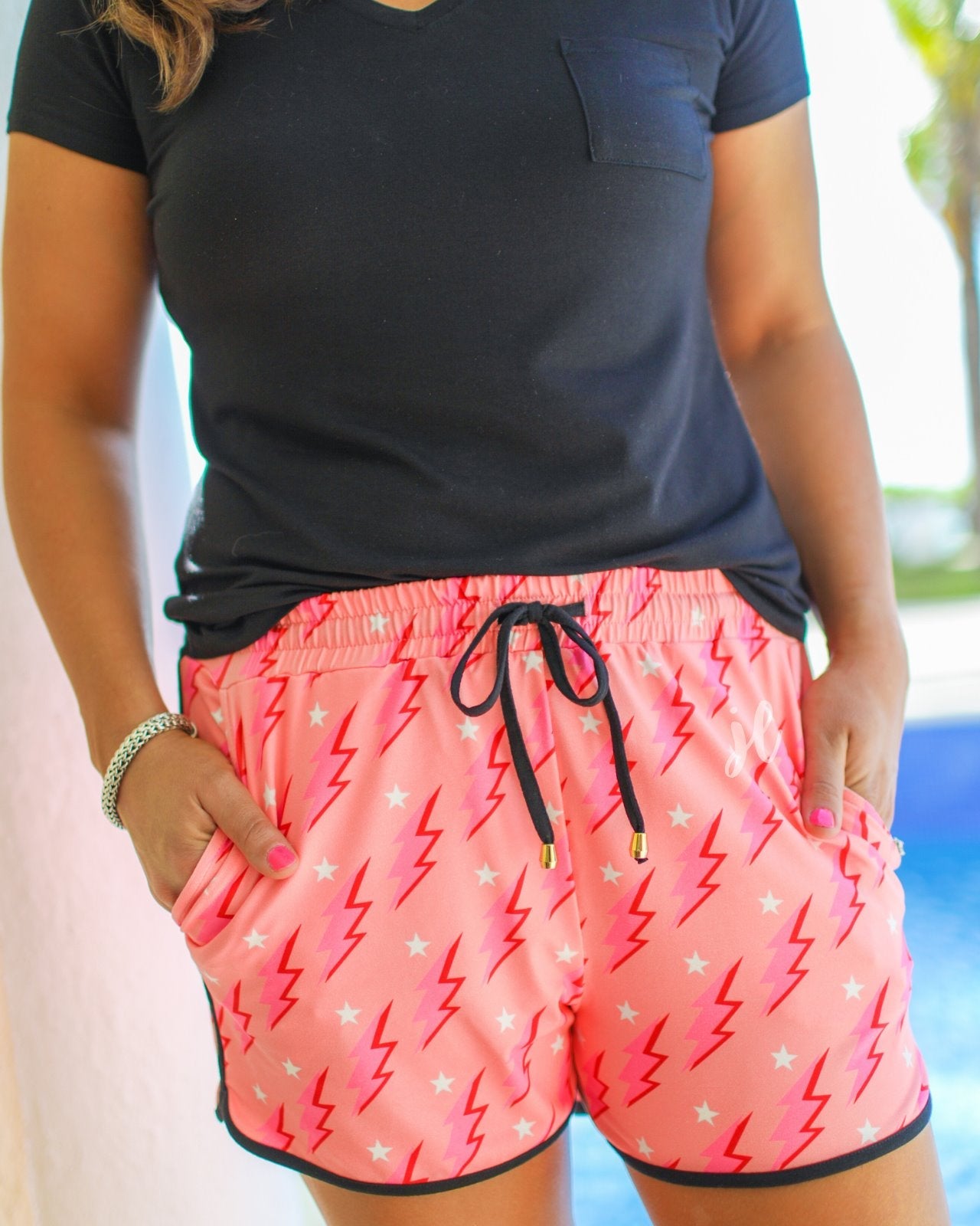 mid-rise relaxed fit activewear shorts with side pockets and a tie waist drawstring with gold bead accents, designed on a two-tone pink and red lightning bolts and white stars pattern print fabric, coral base, and black trim