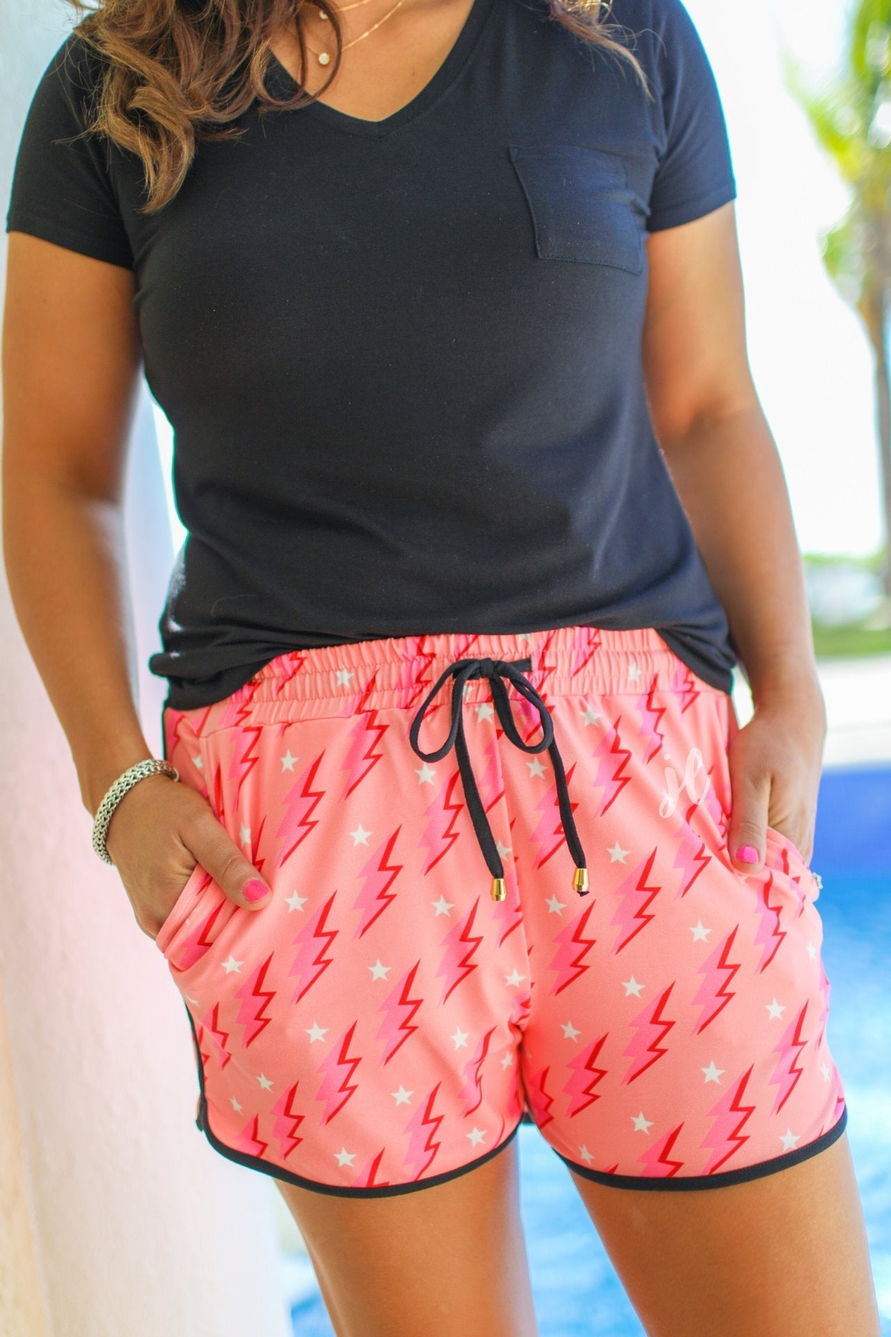 mid-rise relaxed fit activewear shorts with side pockets and a tie waist drawstring with gold bead accents, designed on a two-tone pink and red lightning bolts and white stars pattern print fabric, coral base, and black trim