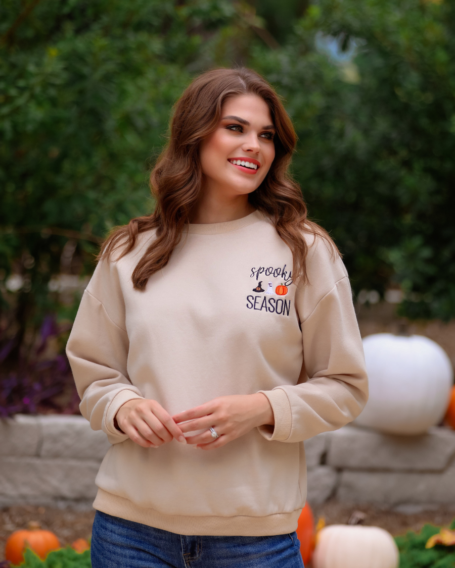 a tan long-sleeve sweatshirt crafted from soft fabric with black embroidered 'spooky SEASON' chest lettering, mini witch hat, ghost, and pumpkin accents, crew neckline, dropped shoulders, ribbed cuffs, banded hem, and a relaxed fit