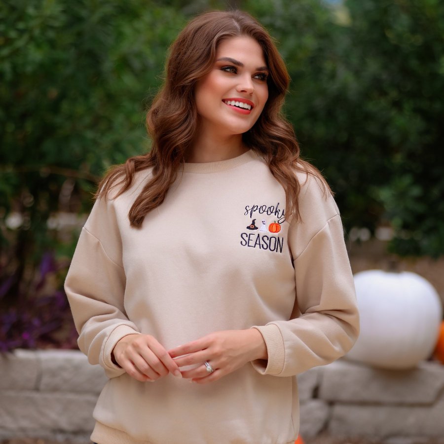 a tan long-sleeve sweatshirt crafted from soft fabric with black embroidered 'spooky SEASON' chest lettering, mini witch hat, ghost, and pumpkin accents, crew neckline, dropped shoulders, ribbed cuffs, banded hem, and a relaxed fit