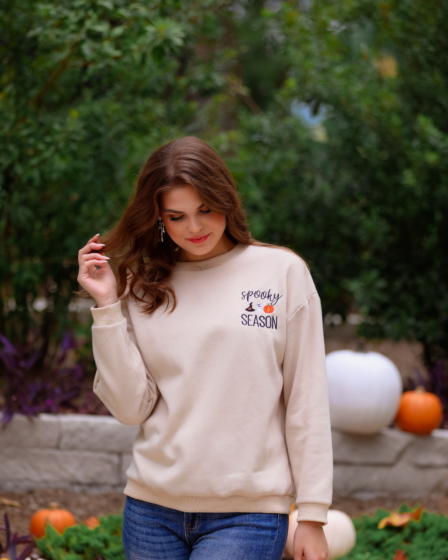 a tan long-sleeve sweatshirt crafted from soft fabric with black embroidered 'spooky SEASON' chest lettering, mini witch hat, ghost, and pumpkin accents, crew neckline, dropped shoulders, ribbed cuffs, banded hem, and a relaxed fit