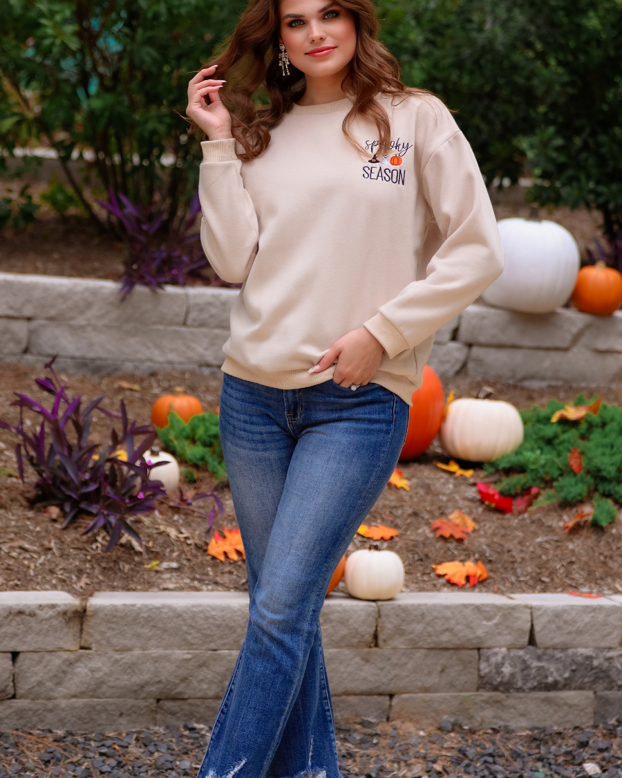 a tan long-sleeve sweatshirt crafted from soft fabric with black embroidered 'spooky SEASON' chest lettering, mini witch hat, ghost, and pumpkin accents, crew neckline, dropped shoulders, ribbed cuffs, banded hem, and a relaxed fit