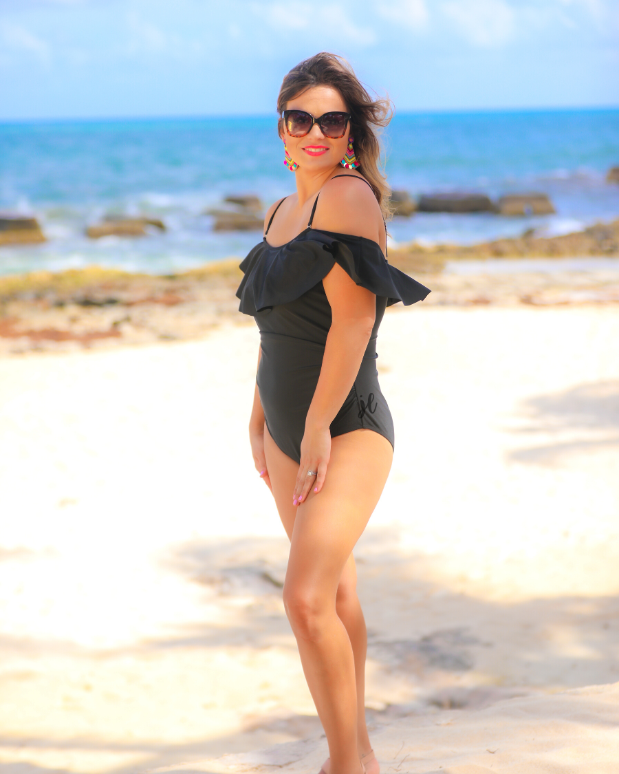 solid black one-piece swimsuit made of stretchy material, a ruffled neckline and straps that can be worn on or off the shoulder, adjustable and removable spaghetti straps, an adjustable crisscrossed cutout back, side ruched detailing, and removable padding