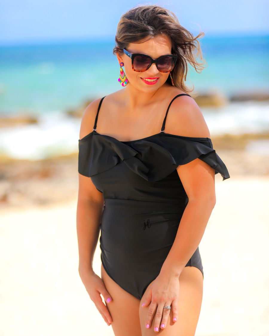 solid black one-piece swimsuit made of stretchy material, a ruffled neckline and straps that can be worn on or off the shoulder, adjustable and removable spaghetti straps, an adjustable crisscrossed cutout back, side ruched detailing, and removable padding