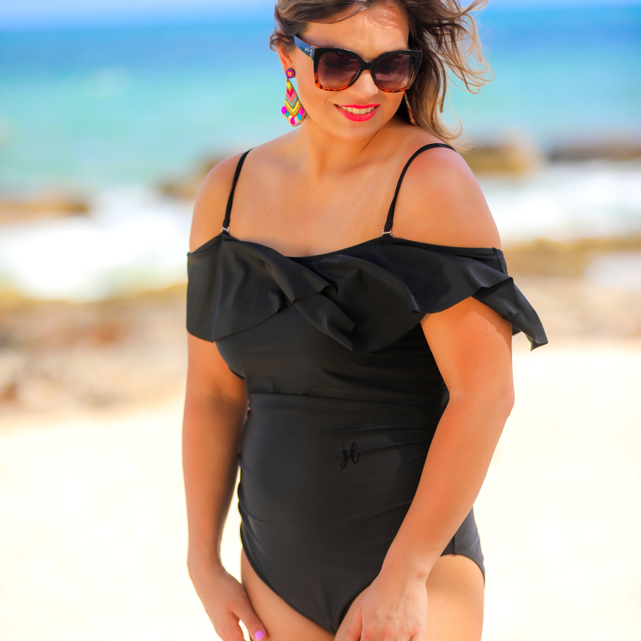 solid black one-piece swimsuit made of stretchy material, a ruffled neckline and straps that can be worn on or off the shoulder, adjustable and removable spaghetti straps, an adjustable crisscrossed cutout back, side ruched detailing, and removable padding