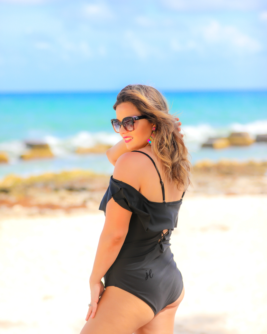 solid black one-piece swimsuit made of stretchy material, a ruffled neckline and straps that can be worn on or off the shoulder, adjustable and removable spaghetti straps, an adjustable crisscrossed cutout back, side ruched detailing, and removable padding