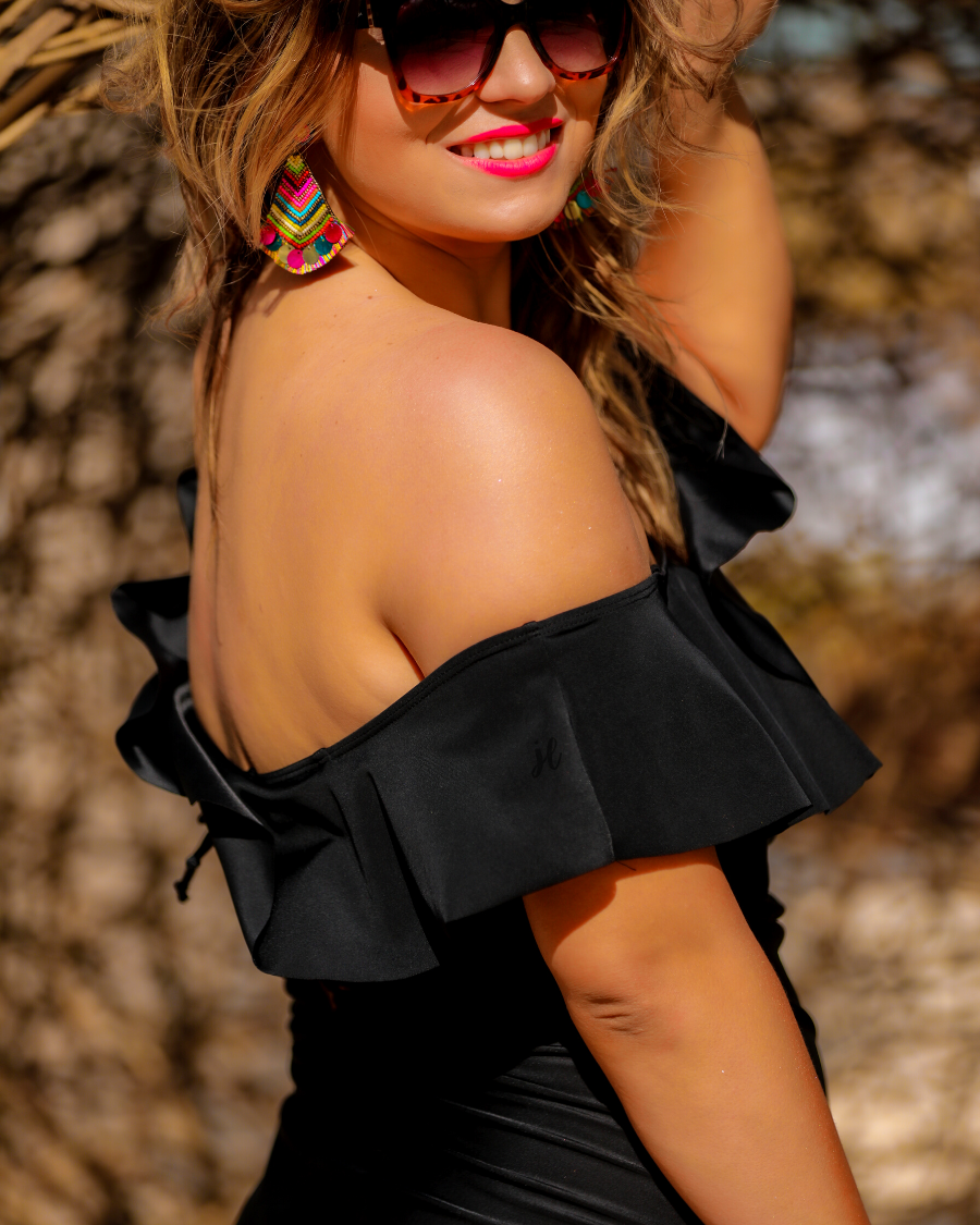 solid black one-piece swimsuit made of stretchy material, a ruffled neckline and straps that can be worn on or off the shoulder, adjustable and removable spaghetti straps, an adjustable crisscrossed cutout back, side ruched detailing, and removable padding