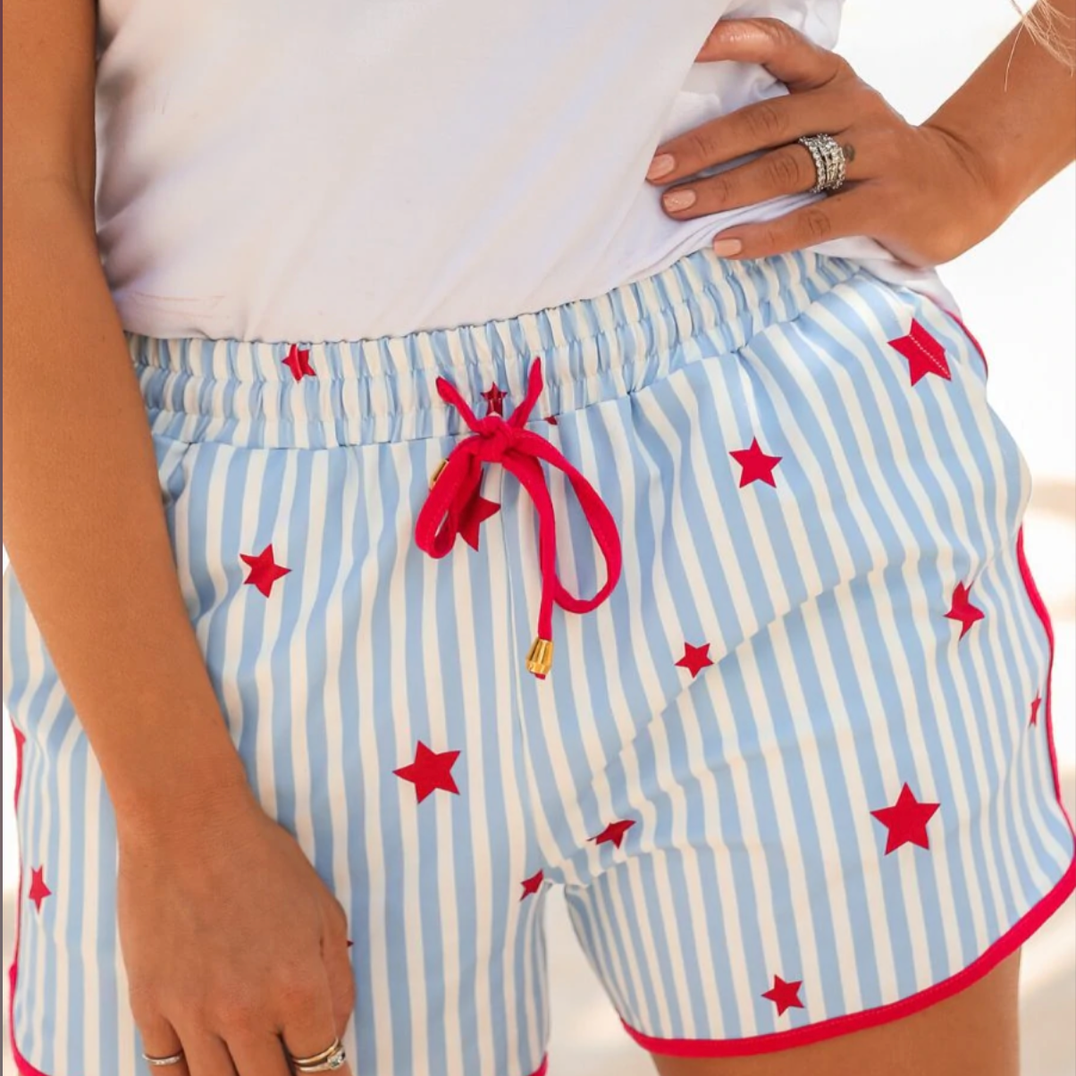 mid-rise relaxed fit activewear shorts with side pockets and a tie waist drawstring with gold bead accents, designed on a mix of blue and white vertical stripes and red stars patterned print fabric and red trim