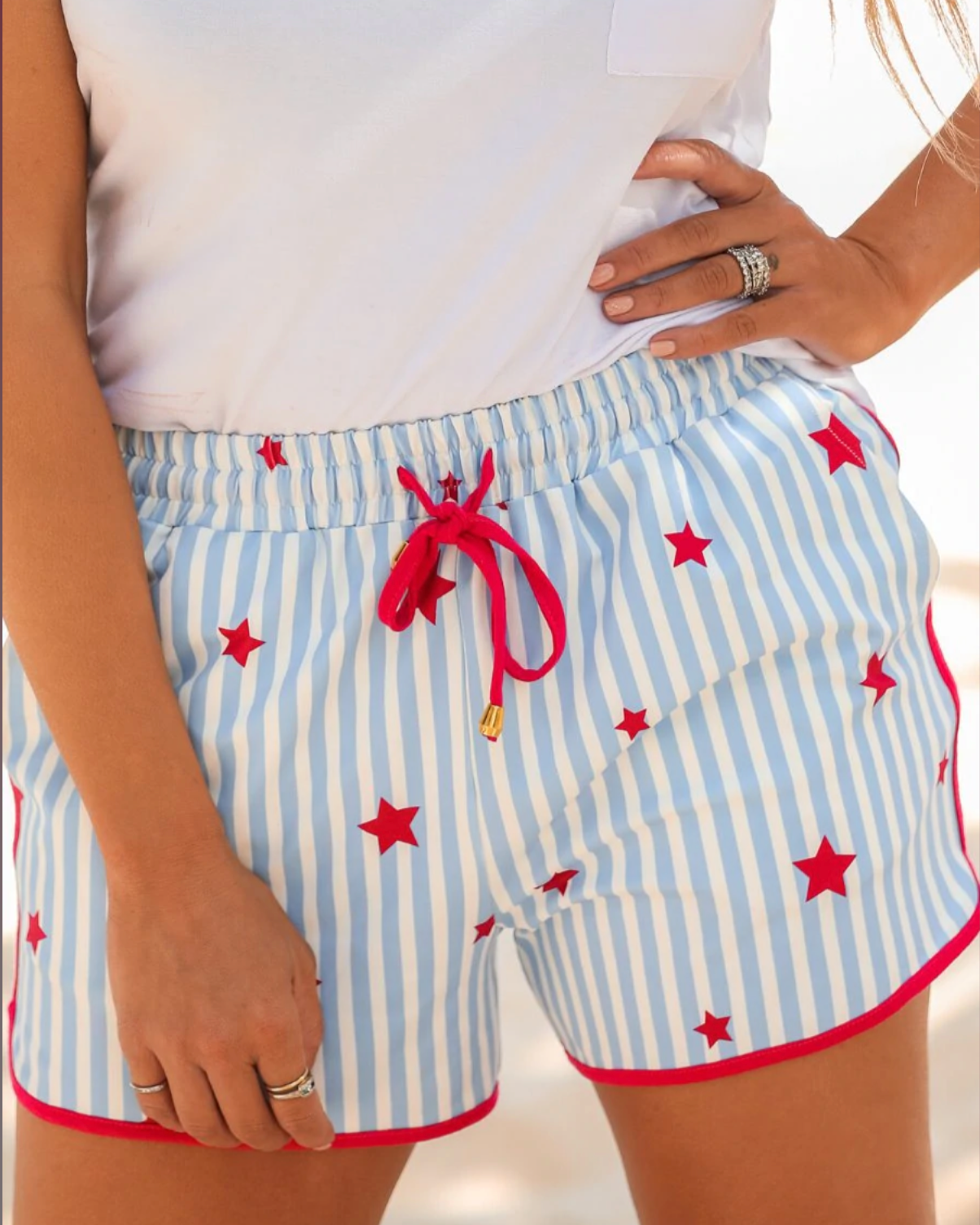 mid-rise relaxed fit activewear shorts with side pockets and a tie waist drawstring with gold bead accents, designed on a mix of blue and white vertical stripes and red stars patterned print fabric and red trim