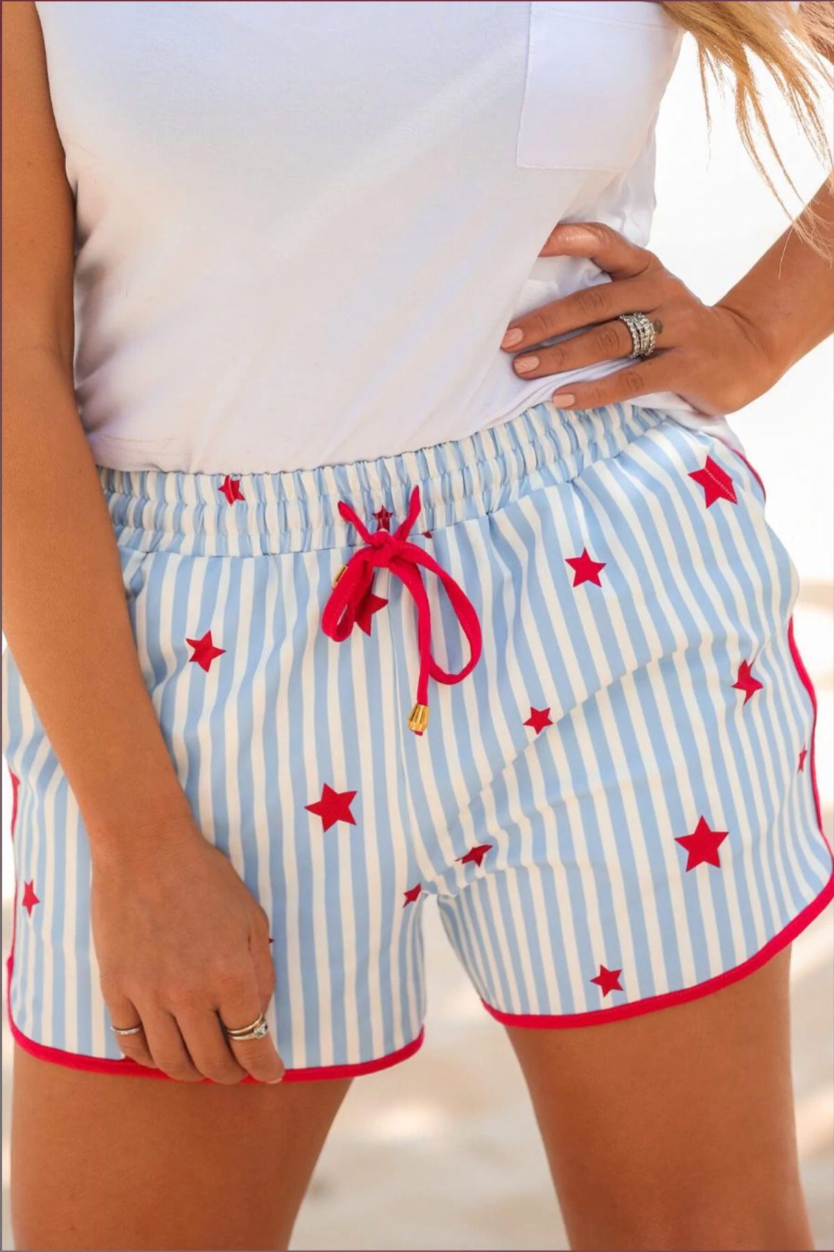 mid-rise relaxed fit activewear shorts with side pockets and a tie waist drawstring with gold bead accents, designed on a mix of blue and white vertical stripes and red stars patterned print fabric and red trim