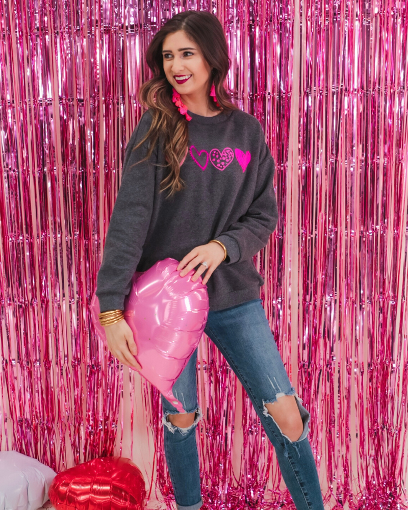a charcoal long-sleeve sweatshirt with a fuzzy inside, sewn-on pink chenille heart patches, a relaxed drop-shoulder fit, and ribbed detailing on the classic crew neckline, cuffs, and hemline