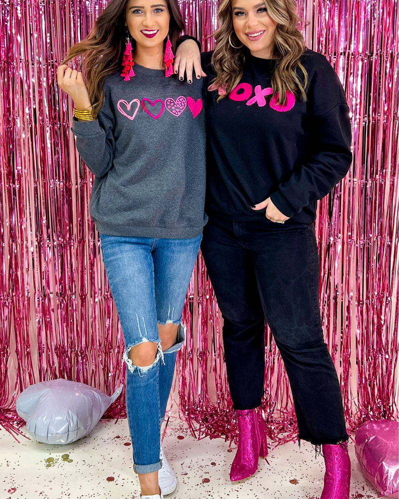 a charcoal long-sleeve sweatshirt with a fuzzy inside, sewn-on pink chenille heart patches, a relaxed drop-shoulder fit, and ribbed detailing on the classic crew neckline, cuffs, and hemline