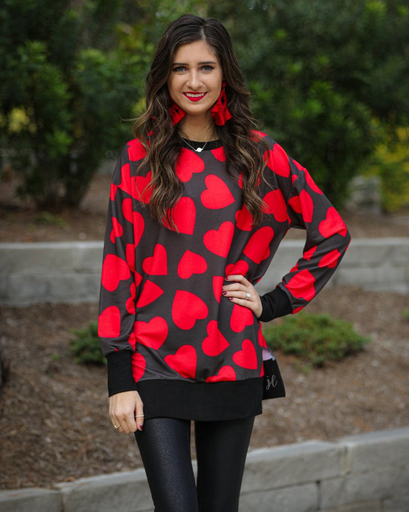 a black long-sleeve tunic with a bold red heart pattern on a black base, side slits, a relaxed drop-shoulder fit, and ribbed detailing on the neckline, cuffs, and hemline