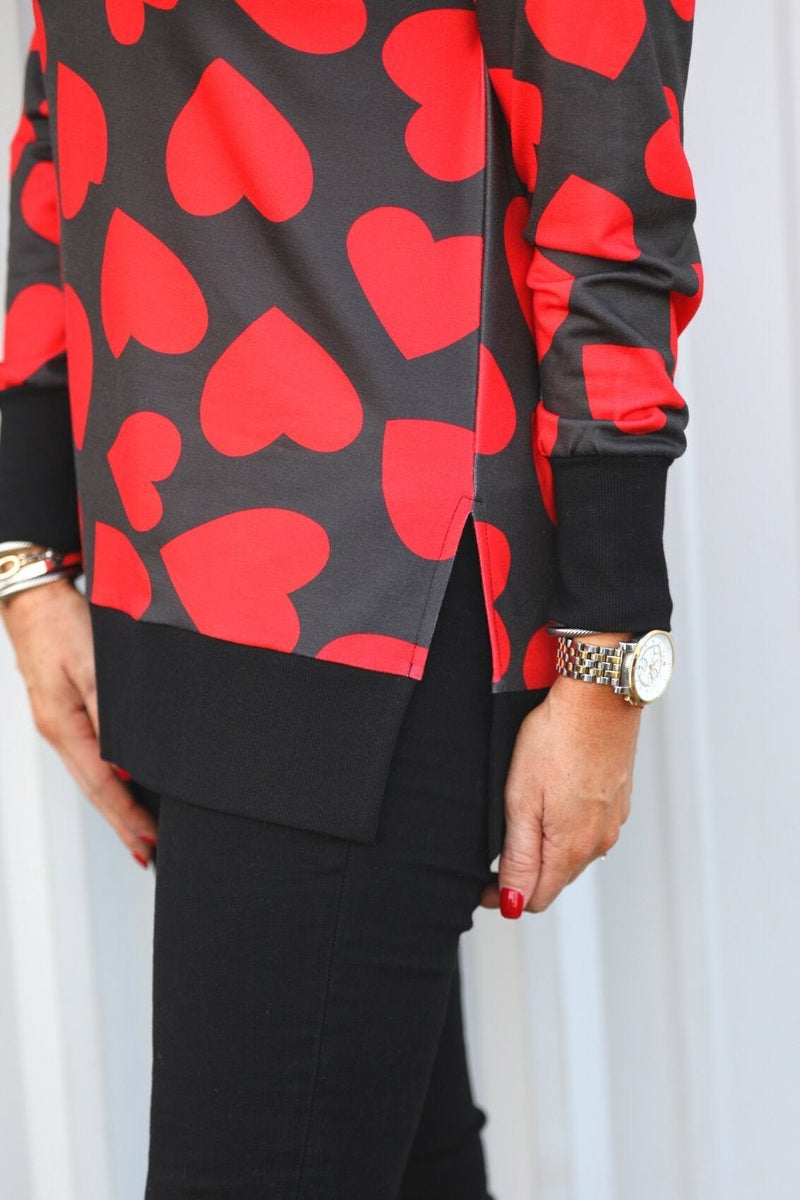 a black long-sleeve tunic with a bold red heart pattern on a black base, side slits, a relaxed drop-shoulder fit, and ribbed detailing on the neckline, cuffs, and hemline