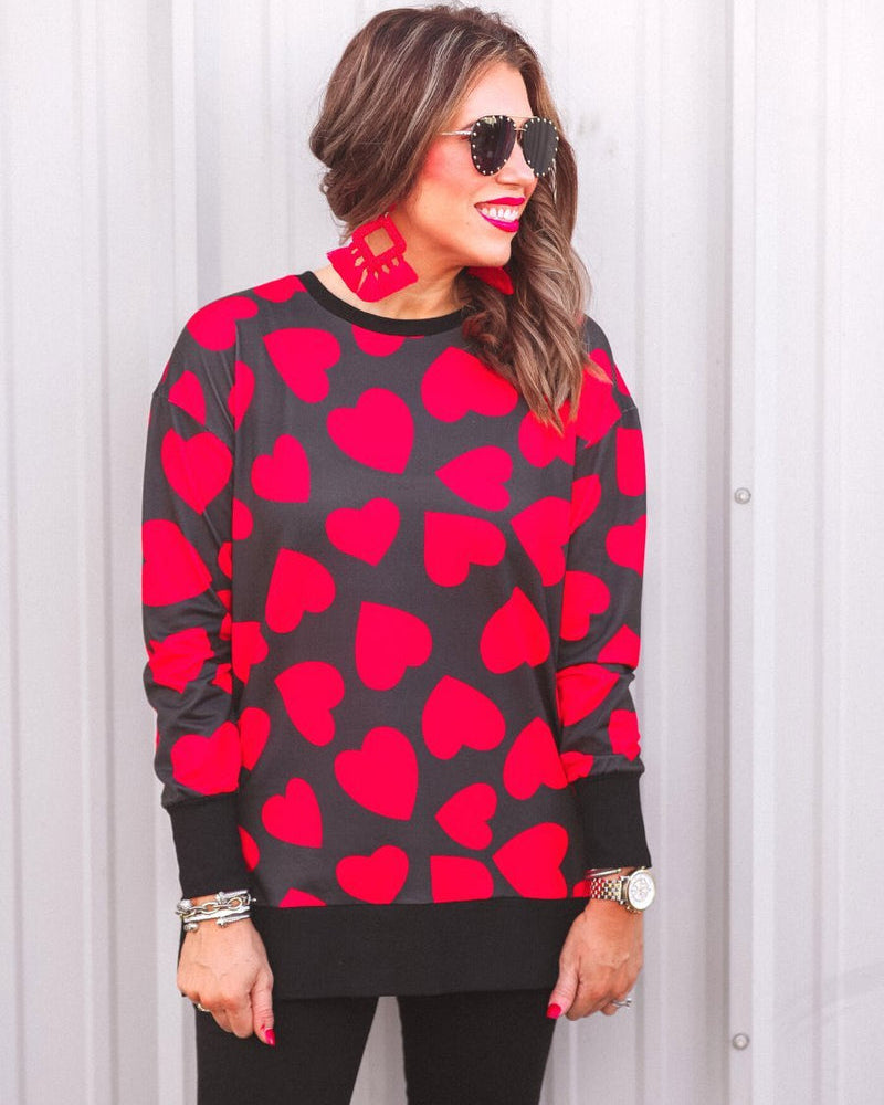 a black long-sleeve tunic with a bold red heart pattern on a black base, side slits, a relaxed drop-shoulder fit, and ribbed detailing on the neckline, cuffs, and hemline