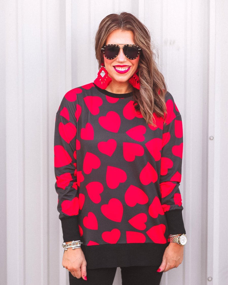 a black long-sleeve tunic with a bold red heart pattern on a black base, side slits, a relaxed drop-shoulder fit, and ribbed detailing on the neckline, cuffs, and hemline