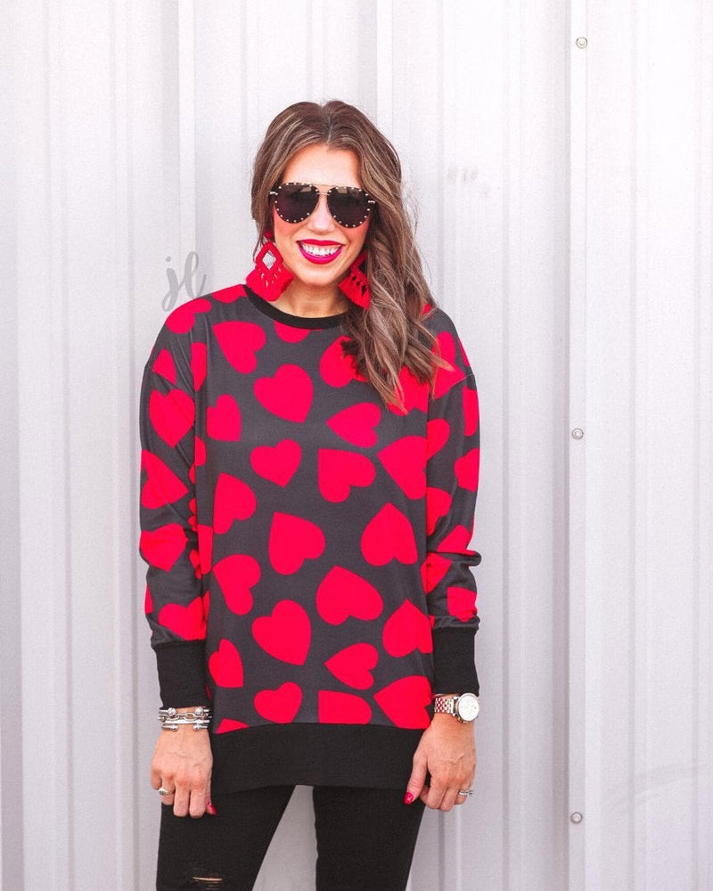 a black long-sleeve tunic with a bold red heart pattern on a black base, side slits, a relaxed drop-shoulder fit, and ribbed detailing on the neckline, cuffs, and hemline