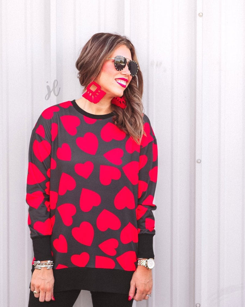 a black long-sleeve tunic with a bold red heart pattern on a black base, side slits, a relaxed drop-shoulder fit, and ribbed detailing on the neckline, cuffs, and hemline