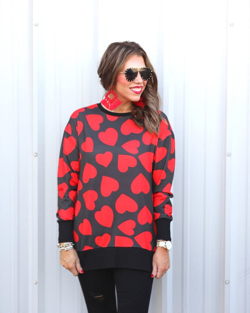a black long-sleeve tunic with a bold red heart pattern on a black base, side slits, a relaxed drop-shoulder fit, and ribbed detailing on the neckline, cuffs, and hemline
