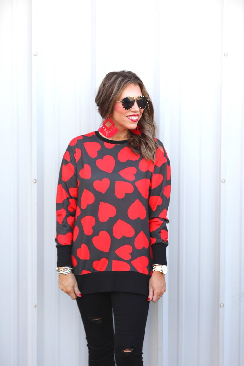a black long-sleeve tunic with a bold red heart pattern on a black base, side slits, a relaxed drop-shoulder fit, and ribbed detailing on the neckline, cuffs, and hemline