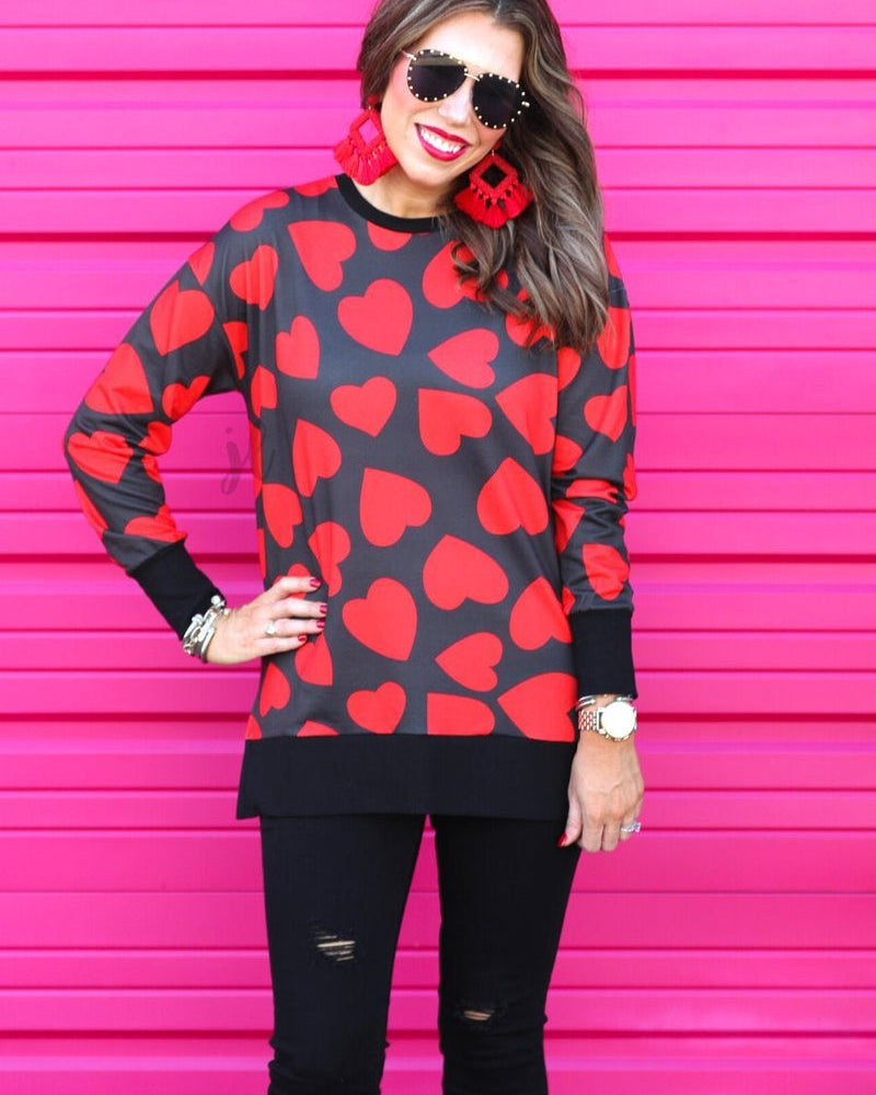 a black long-sleeve tunic with a bold red heart pattern on a black base, side slits, a relaxed drop-shoulder fit, and ribbed detailing on the neckline, cuffs, and hemline