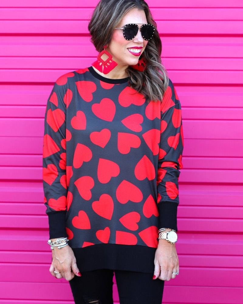 a black long-sleeve tunic with a bold red heart pattern on a black base, side slits, a relaxed drop-shoulder fit, and ribbed detailing on the neckline, cuffs, and hemline
