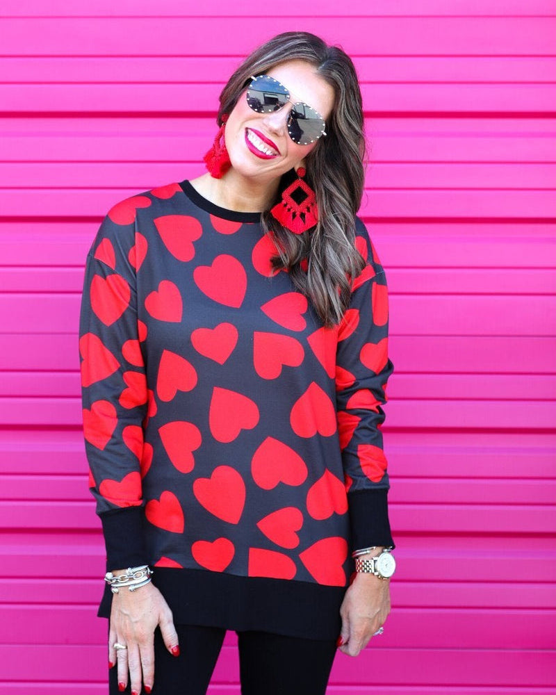 a black long-sleeve tunic with a bold red heart pattern on a black base, side slits, a relaxed drop-shoulder fit, and ribbed detailing on the neckline, cuffs, and hemline