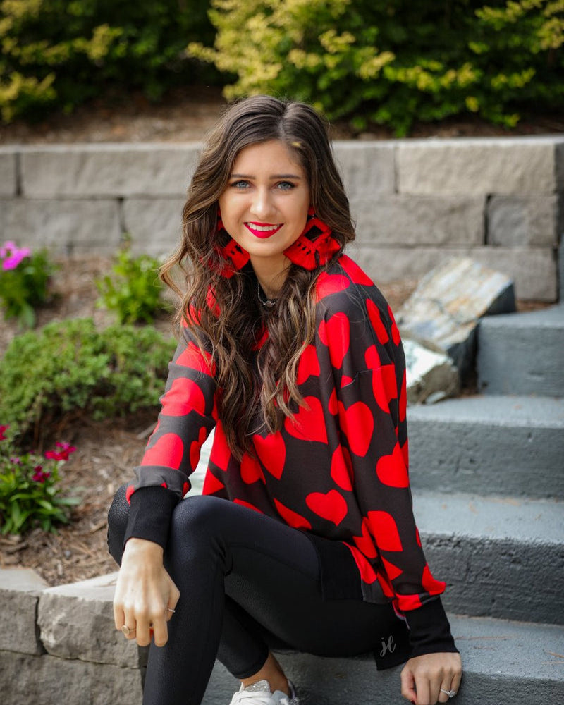 a black long-sleeve tunic with a bold red heart pattern on a black base, side slits, a relaxed drop-shoulder fit, and ribbed detailing on the neckline, cuffs, and hemline