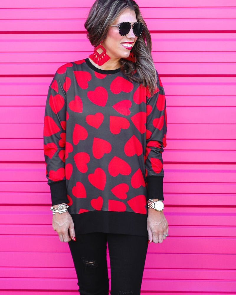 a black long-sleeve tunic with a bold red heart pattern on a black base, side slits, a relaxed drop-shoulder fit, and ribbed detailing on the neckline, cuffs, and hemline
