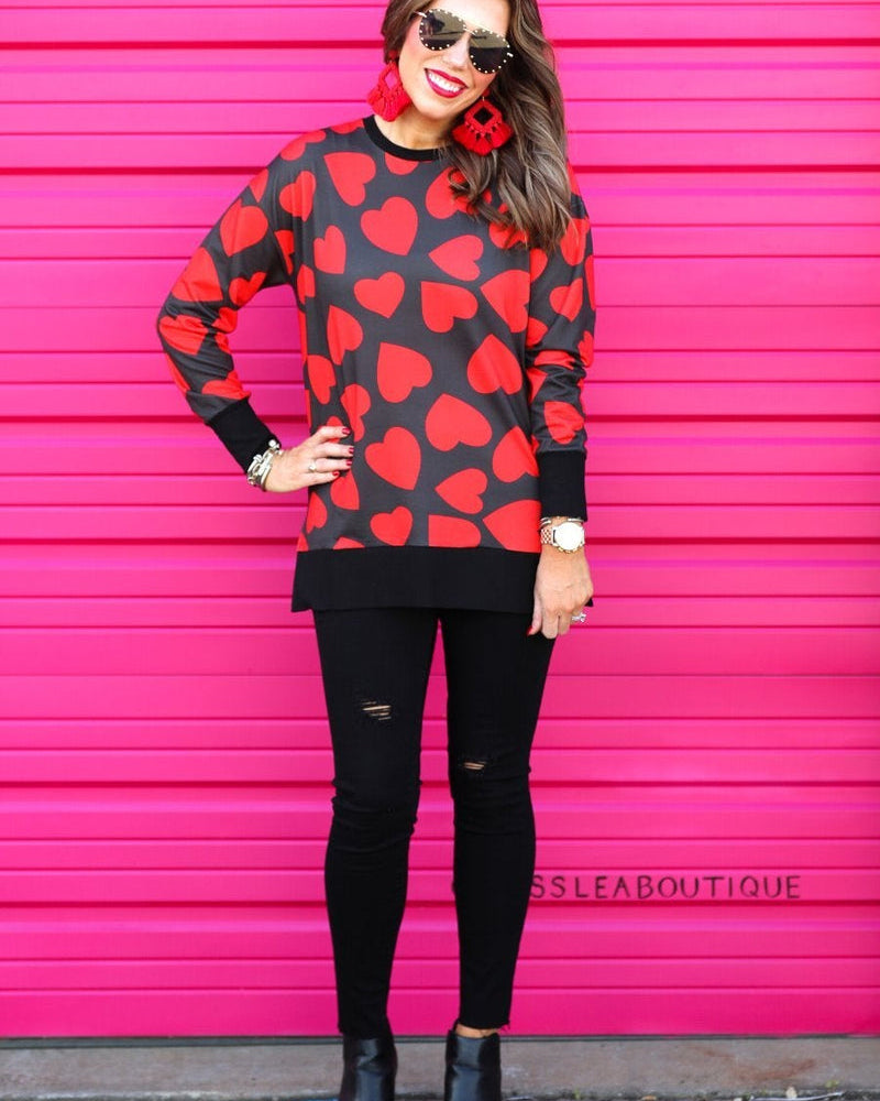 a black long-sleeve tunic with a bold red heart pattern on a black base, side slits, a relaxed drop-shoulder fit, and ribbed detailing on the neckline, cuffs, and hemline