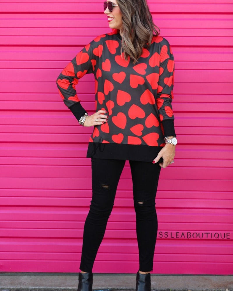 a black long-sleeve tunic with a bold red heart pattern on a black base, side slits, a relaxed drop-shoulder fit, and ribbed detailing on the neckline, cuffs, and hemline