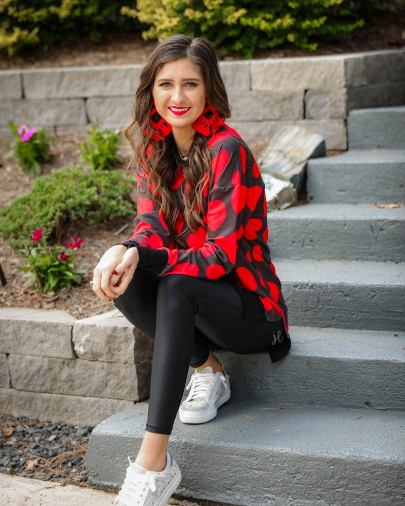 a black long-sleeve tunic with a bold red heart pattern on a black base, side slits, a relaxed drop-shoulder fit, and ribbed detailing on the neckline, cuffs, and hemline