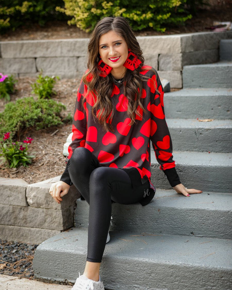 a black long-sleeve tunic with a bold red heart pattern on a black base, side slits, a relaxed drop-shoulder fit, and ribbed detailing on the neckline, cuffs, and hemline