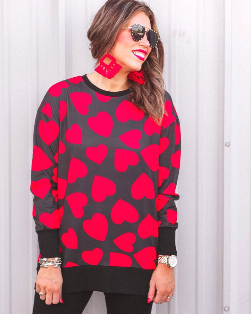 a black long-sleeve tunic with a bold red heart pattern on a black base, side slits, a relaxed drop-shoulder fit, and ribbed detailing on the neckline, cuffs, and hemline