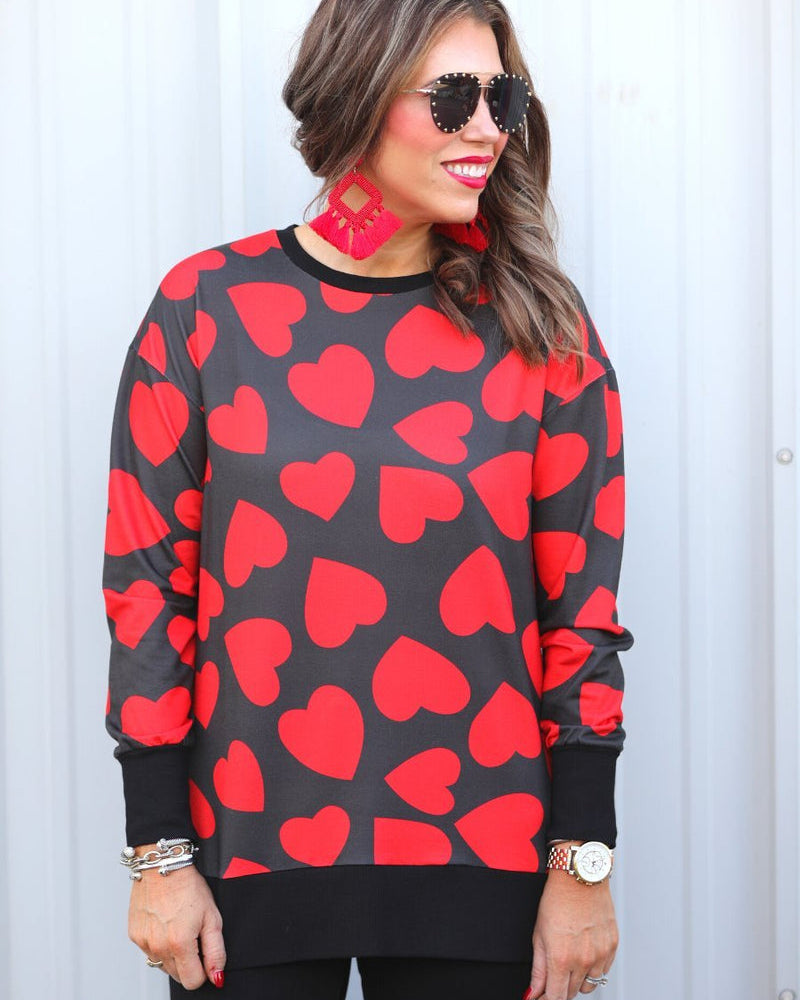 a black long-sleeve tunic with a bold red heart pattern on a black base, side slits, a relaxed drop-shoulder fit, and ribbed detailing on the neckline, cuffs, and hemline