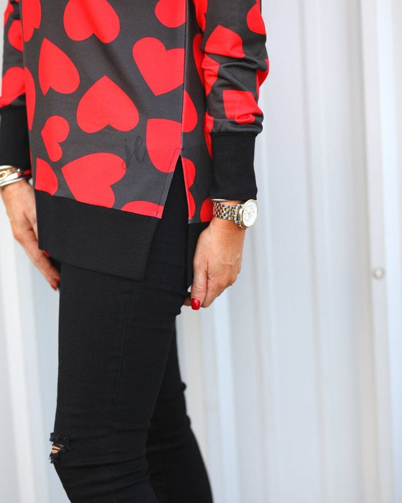 a black long-sleeve tunic with a bold red heart pattern on a black base, side slits, a relaxed drop-shoulder fit, and ribbed detailing on the neckline, cuffs, and hemline