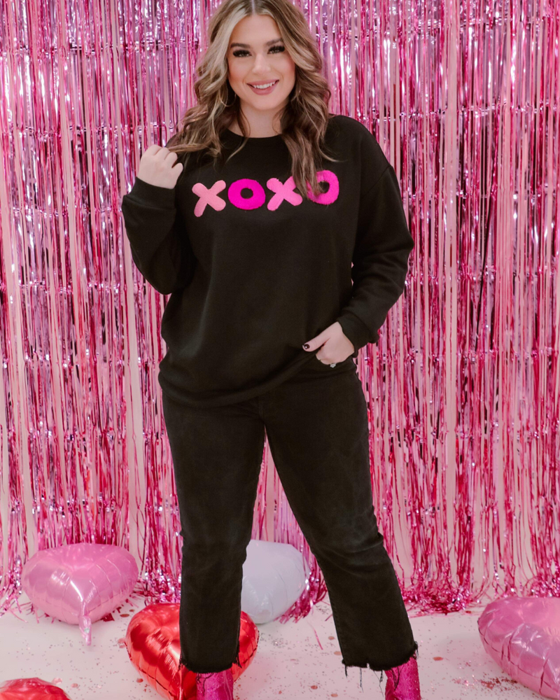 a black long-sleeve sweatshirt with a soft fleece interior, a drop-shoulder silhouette, ribbed detailing on the classic crew neckline, cuffs, and hemline, finished with bold pink sewn-on chenille 'XOXO' patches
