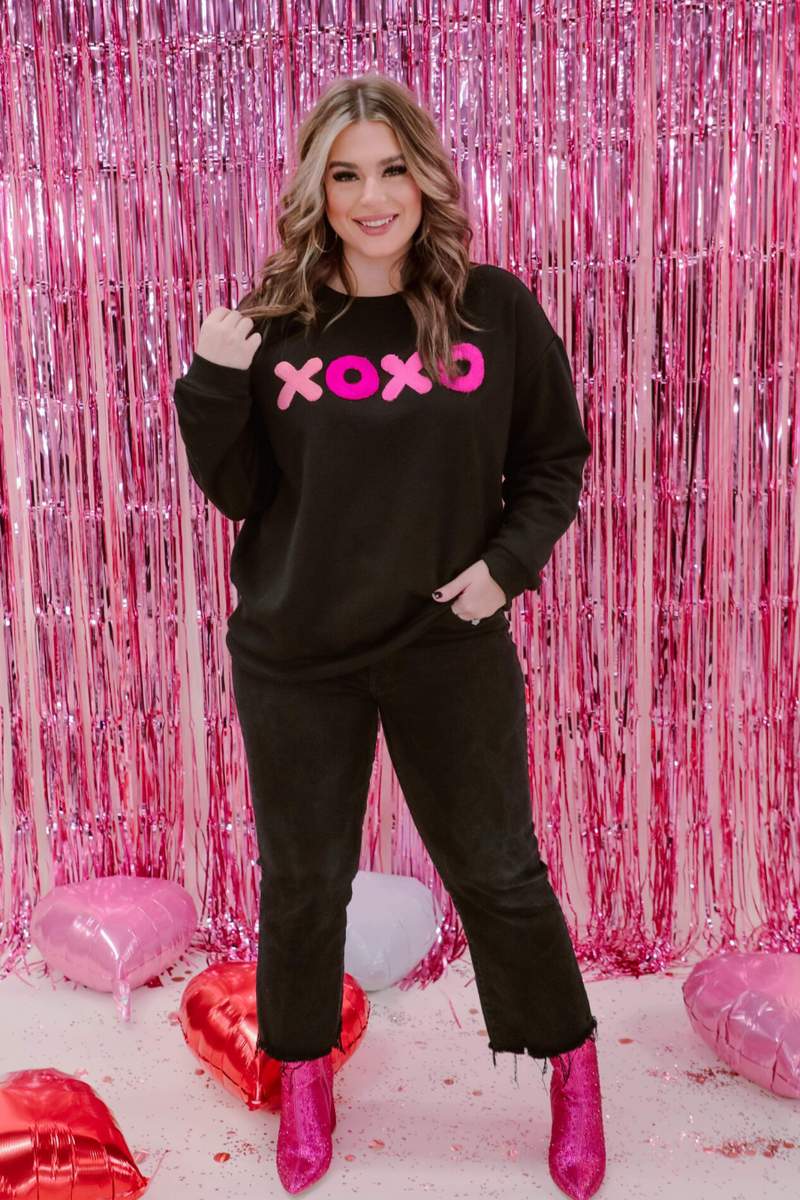 a black long-sleeve sweatshirt with a soft fleece interior, a drop-shoulder silhouette, ribbed detailing on the classic crew neckline, cuffs, and hemline, finished with bold pink sewn-on chenille 'XOXO' patches