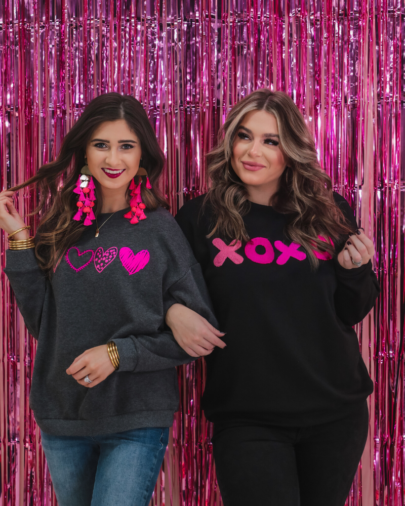 a black long-sleeve sweatshirt with a soft fleece interior, a drop-shoulder silhouette, ribbed detailing on the classic crew neckline, cuffs, and hemline, finished with bold pink sewn-on chenille 'XOXO' patches