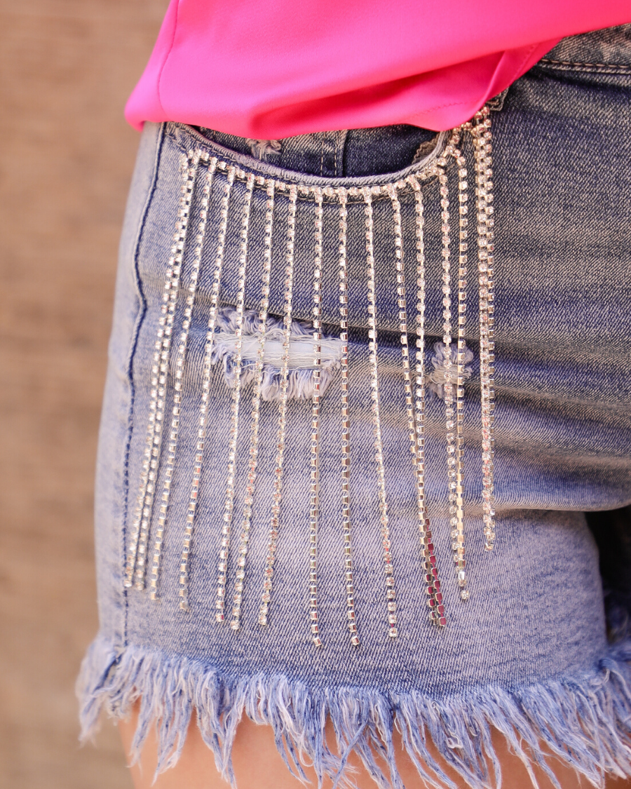 mid-rise denim shorts with rhinestone fringe, a frayed hem, full classic pockets, button zip fly with belt loops, distressed detailing, and a medium wash